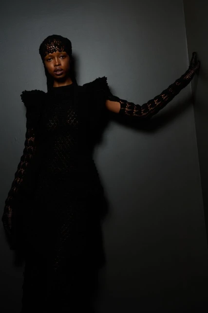 Portrait of a Black woman in a black lace dress and long gloves, posing against a dark gray wall in a fashion studio.