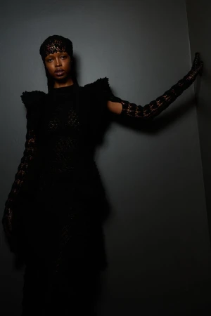Portrait of a Black woman in a black lace dress and long gloves, posing against a dark gray wall in a fashion studio.