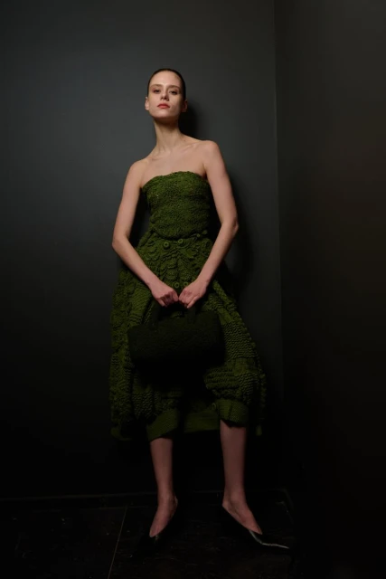 A slender fashion model in a green textured strapless dress stands in a studio against a gray wall, holding a matching green handbag.