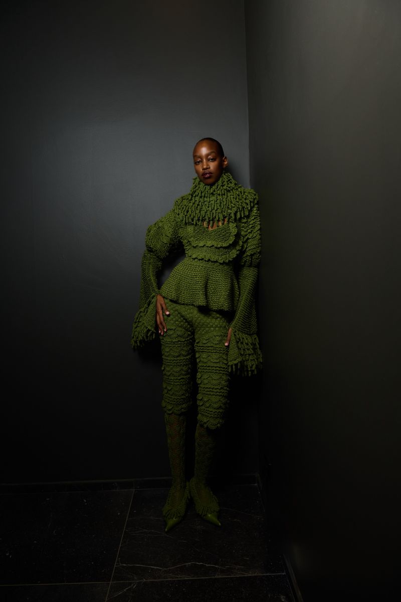 Woman in a green textured pantsuit with a peplum top and bell sleeves poses in front of a grey wall with a dark marble floor.
