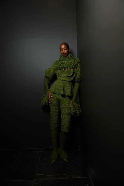 Woman in a green textured pantsuit with a peplum top and bell sleeves poses in front of a grey wall with a dark marble floor.