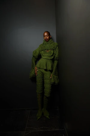 Woman in a green textured pantsuit with a peplum top and bell sleeves poses in front of a grey wall with a dark marble floor.