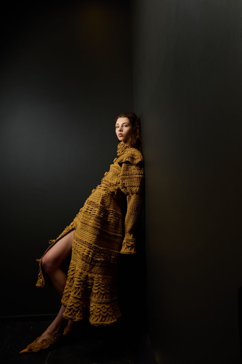 A woman in a distinctive crocheted golden-brown gown poses dramatically against a dark wall, accentuated by directional lighting.