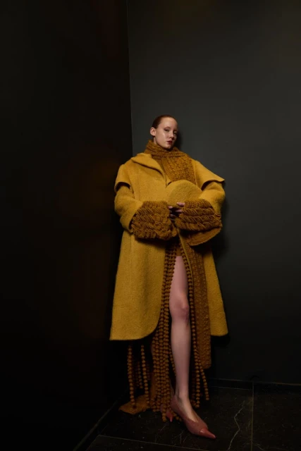Fashionable woman in mustard-yellow coat, scarf, and shoes poses in a corner with a dark background, showcasing unique knitted textures.