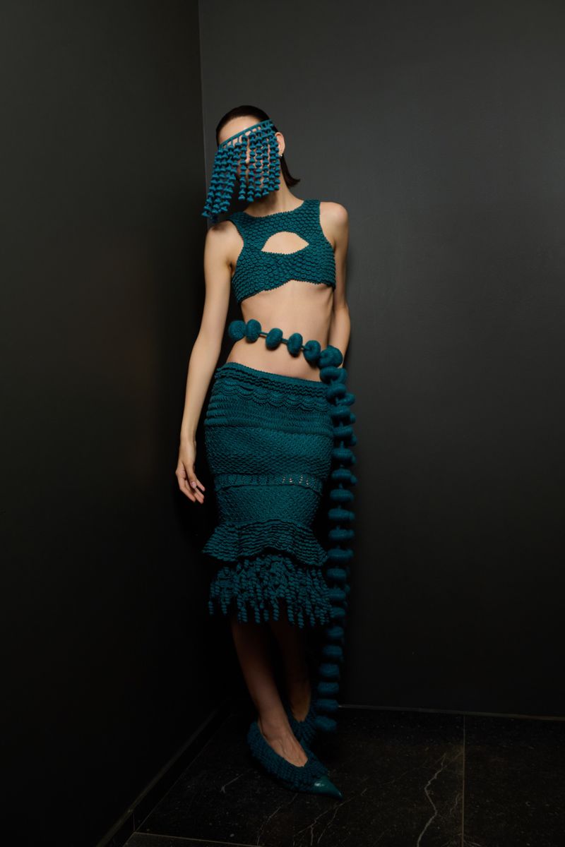 Woman in knitwear two-piece set featuring a cropped top, midi skirt, and matching mask, standing in front of a dark gray corner.