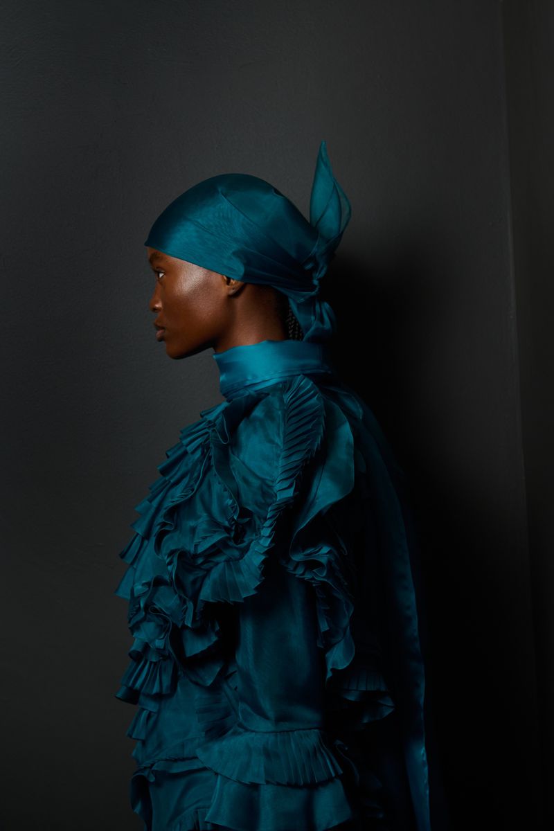 Profile shot of a woman with dark skin wearing a teal ruffled dress and headscarf, posing against a dark gray backdrop, studio lighting.