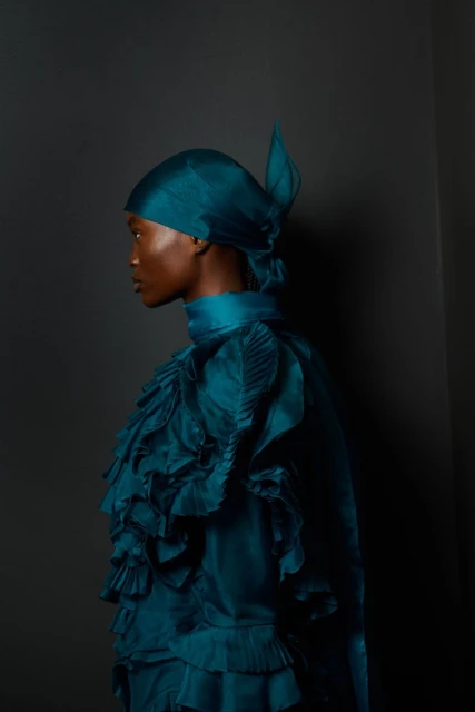 Profile shot of a woman with dark skin wearing a teal ruffled dress and headscarf, posing against a dark gray backdrop, studio lighting.