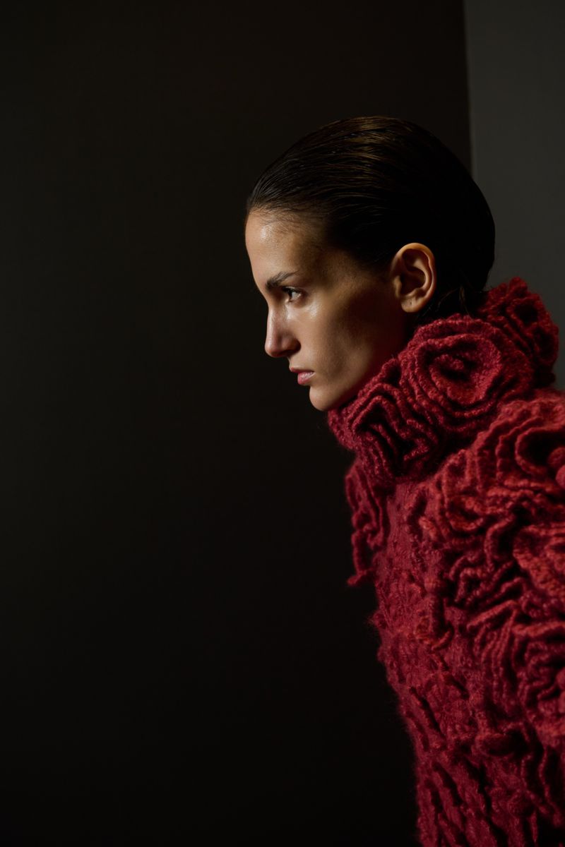 Close-up profile portrait of an adult woman with slicked-back hair wearing a red textured high-neck sweater against a dark background.