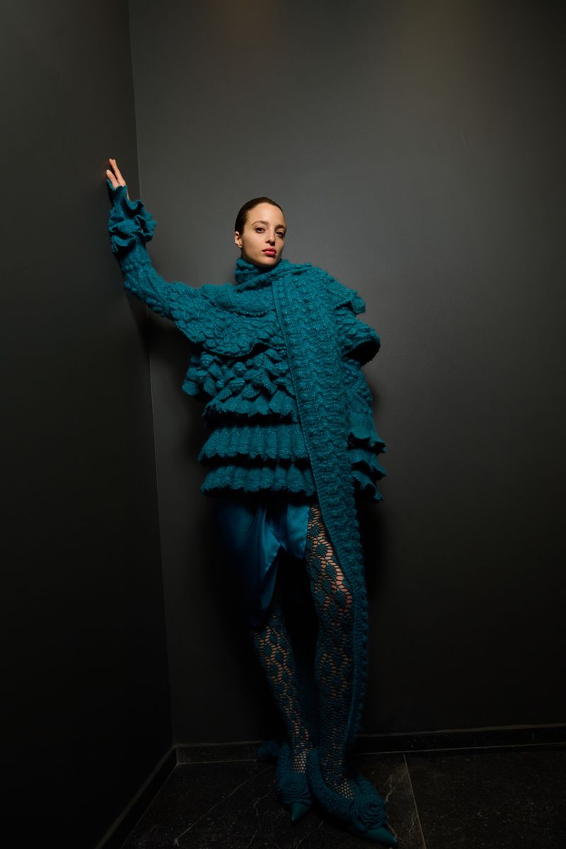 A stylish adult female with fair skin, wearing a turquoise dress with lace tights posing in front of a dark grey wall.