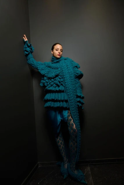 A stylish adult female with fair skin, wearing a turquoise dress with lace tights posing in front of a dark grey wall.