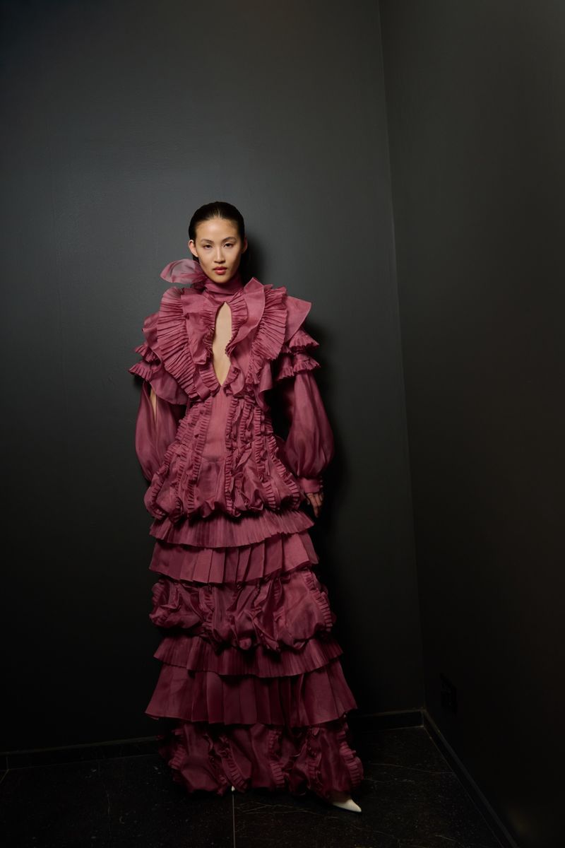 Woman poses in an elaborate, floor-length, mauve gown with ruffles against a dark wall. Indoor fashion shot with dramatic lighting.