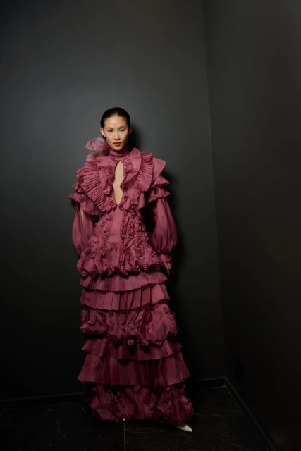 Woman poses in an elaborate, floor-length, mauve gown with ruffles against a dark wall. Indoor fashion shot with dramatic lighting.