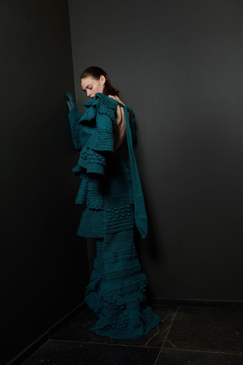 Fashion shot of a woman in a turquoise tiered knit dress posing against a dark grey corner in a minimalist, high-fashion studio setting.
