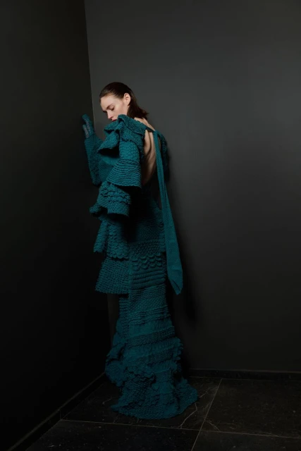 Fashion shot of a woman in a turquoise tiered knit dress posing against a dark grey corner in a minimalist, high-fashion studio setting.