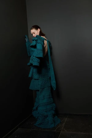 Fashion shot of a woman in a turquoise tiered knit dress posing against a dark grey corner in a minimalist, high-fashion studio setting.