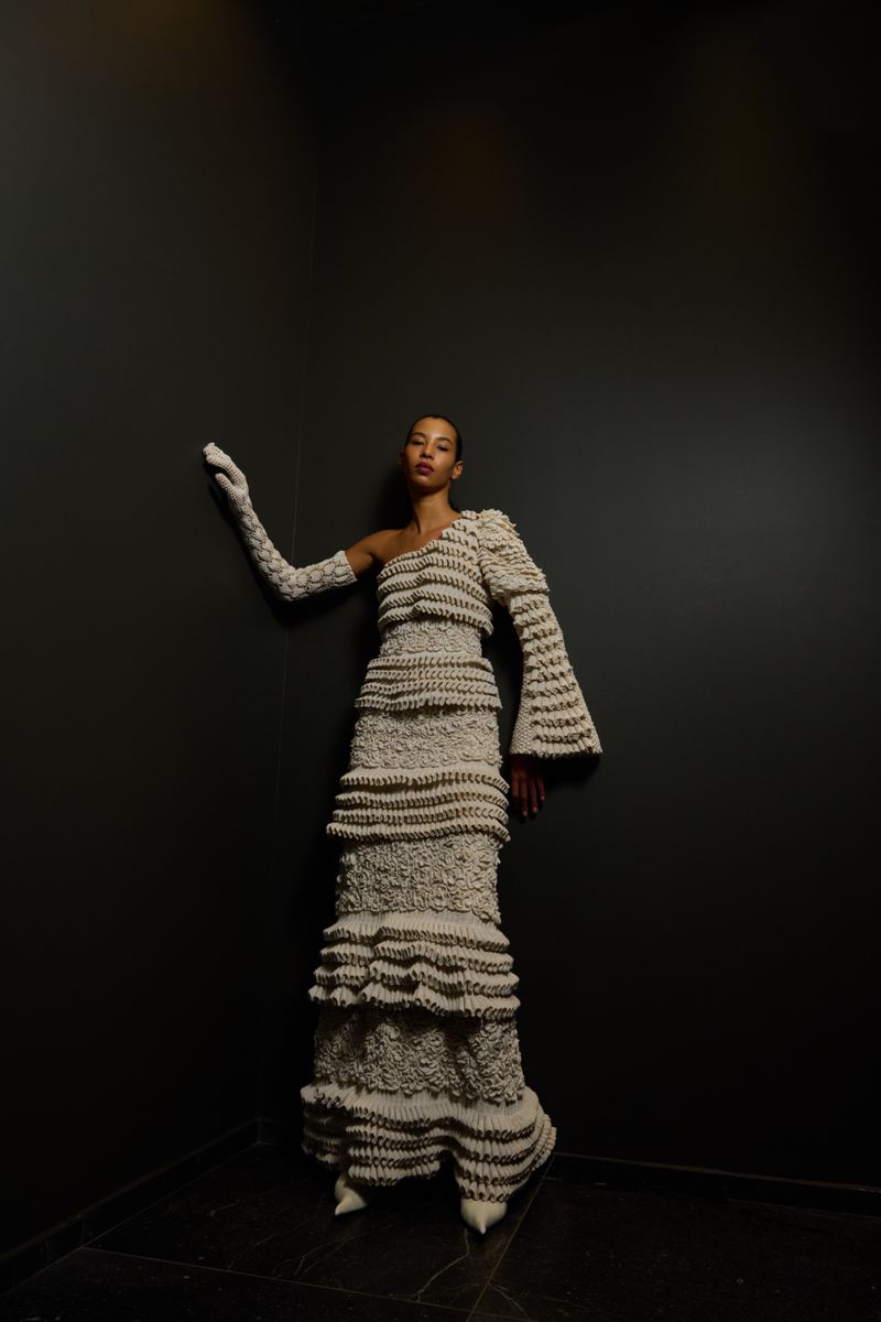 Elegant woman in a textured beige one-shoulder gown and long gloves poses against a dark background in a fashion editorial.
