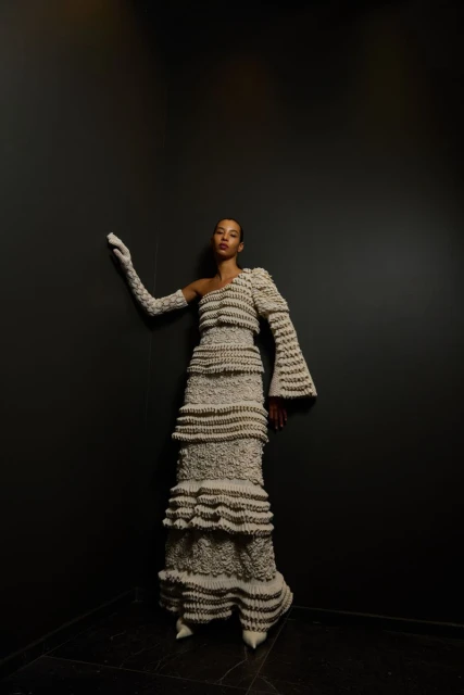 Elegant woman in a textured beige one-shoulder gown and long gloves poses against a dark background in a fashion editorial.