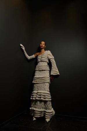 Elegant woman in a textured beige one-shoulder gown and long gloves poses against a dark background in a fashion editorial.