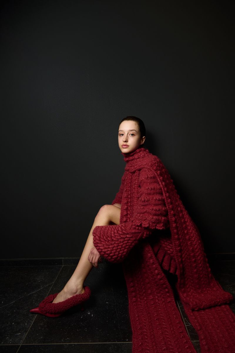 A slender woman with dark hair models a red, textured knitwear outfit with matching shoes against a dark grey studio backdrop, fashion forward.