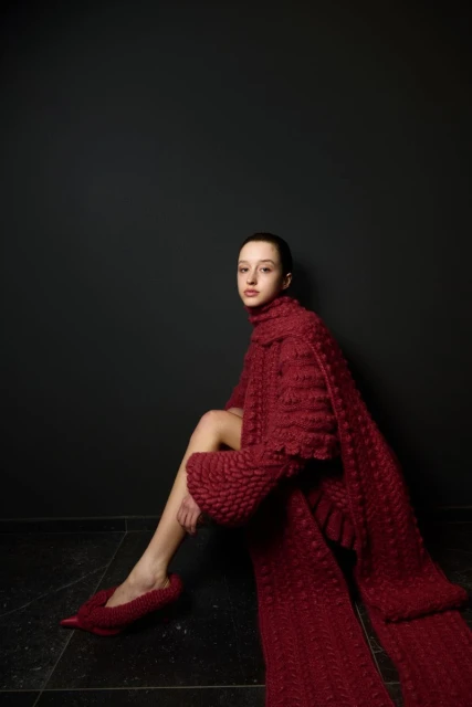 A slender woman with dark hair models a red, textured knitwear outfit with matching shoes against a dark grey studio backdrop, fashion forward.