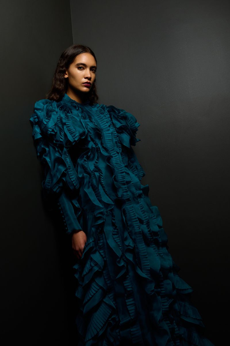 Dark-haired woman in a teal gown with ruffles poses against a dark corner, looking at the camera in a studio setting.