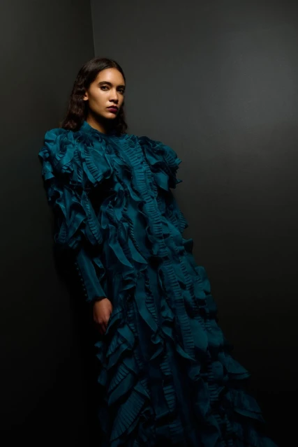 Dark-haired woman in a teal gown with ruffles poses against a dark corner, looking at the camera in a studio setting.