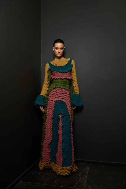 A woman with her hair slicked back poses in an indoor studio wearing a floor-length gown with mustard yellow, pink, green, and teal colors.