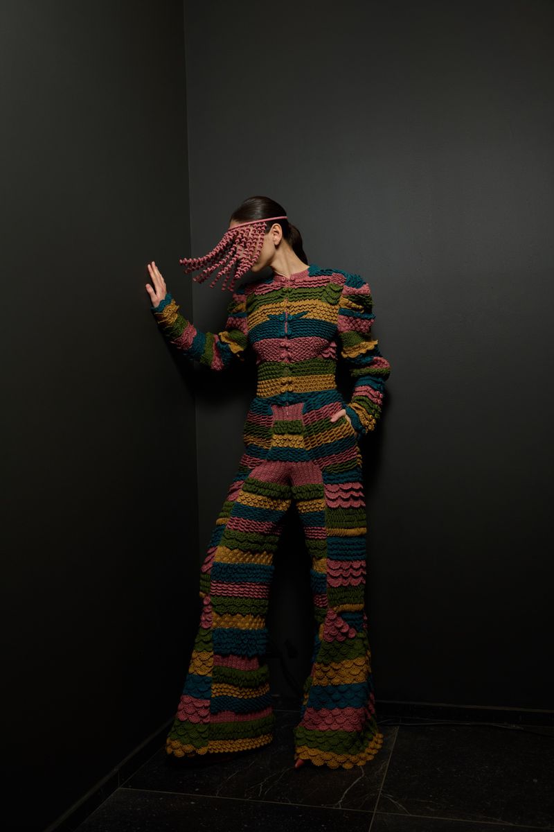 A person wearing a multicolored, striped knit jumpsuit and matching head covering leans against a dark gray corner in a studio setting.