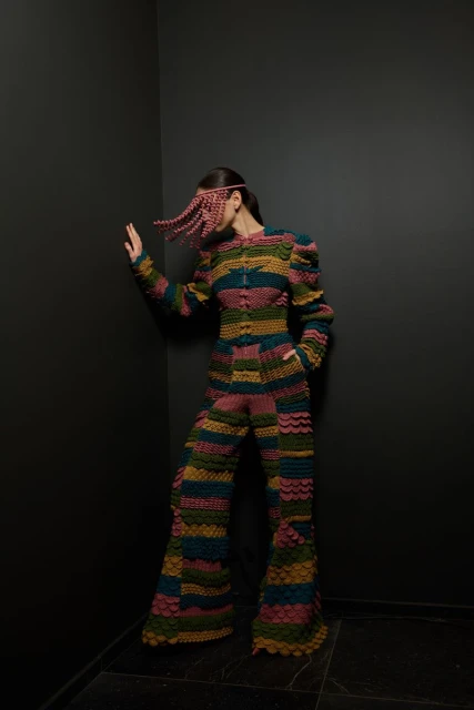 A person wearing a multicolored, striped knit jumpsuit and matching head covering leans against a dark gray corner in a studio setting.