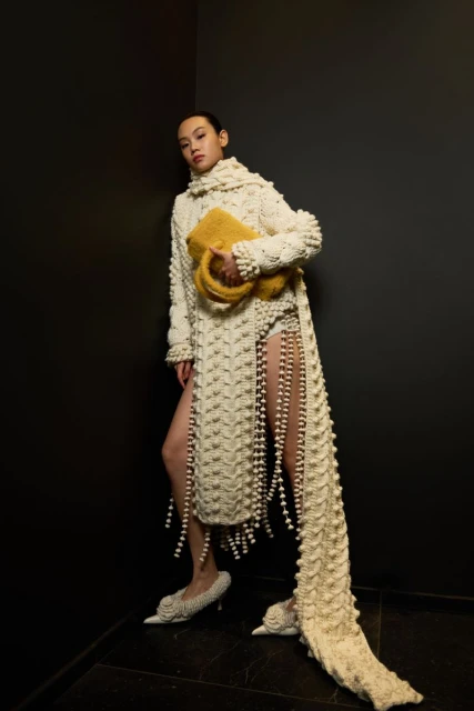 Woman in a knitted, high-necked, floor-length sweater dress with beaded tassels, patterned heels, and yellow bag poses against a black corner.