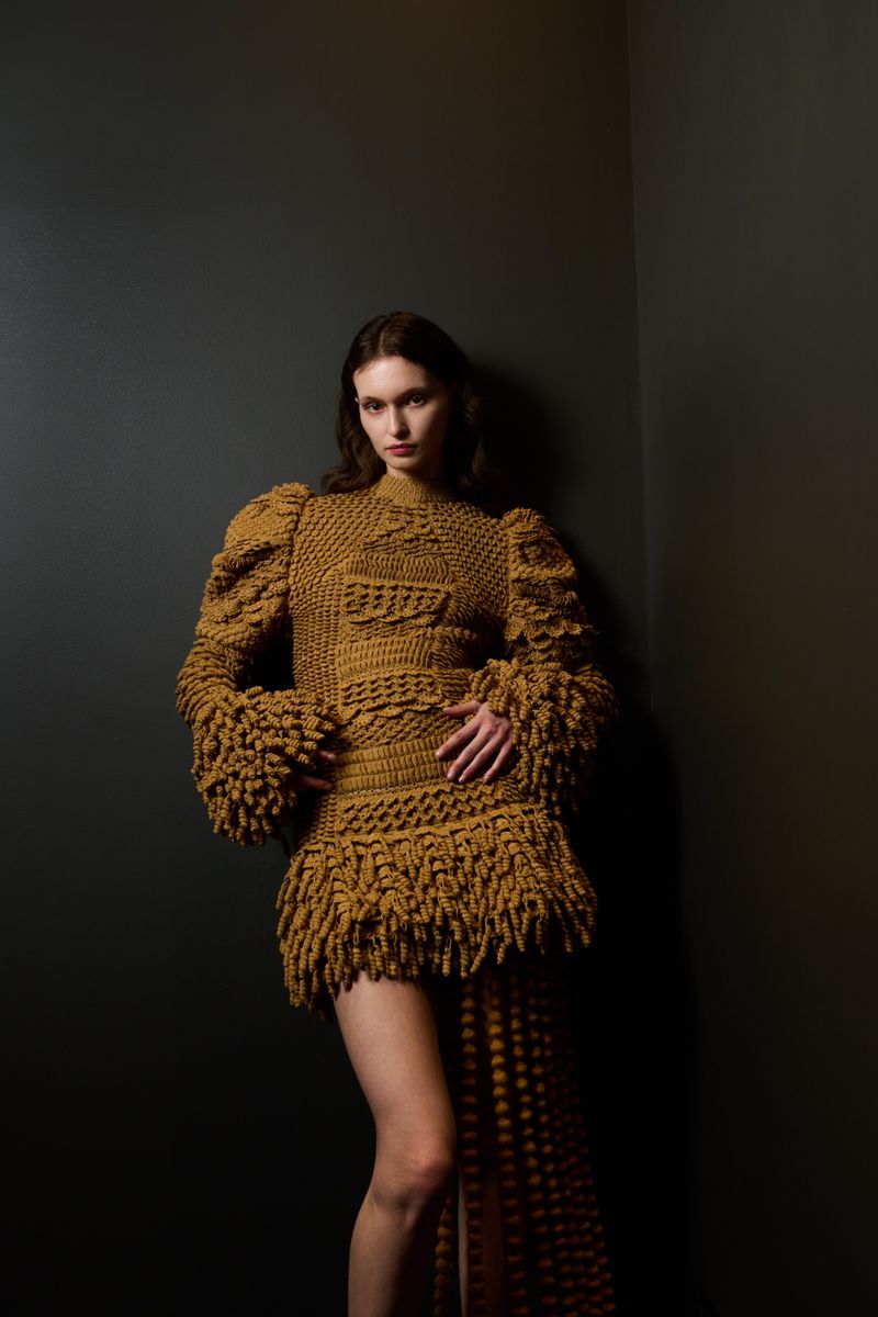 Woman poses in a textured yellow-brown mini-dress with fringe and puff sleeves against a dark gray corner in a studio setting.