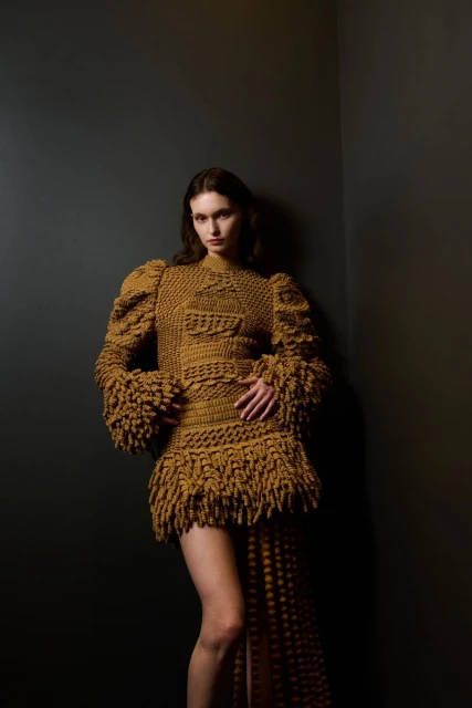 Woman poses in a textured yellow-brown mini-dress with fringe and puff sleeves against a dark gray corner in a studio setting.