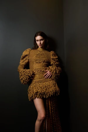 Woman poses in a textured yellow-brown mini-dress with fringe and puff sleeves against a dark gray corner in a studio setting.
