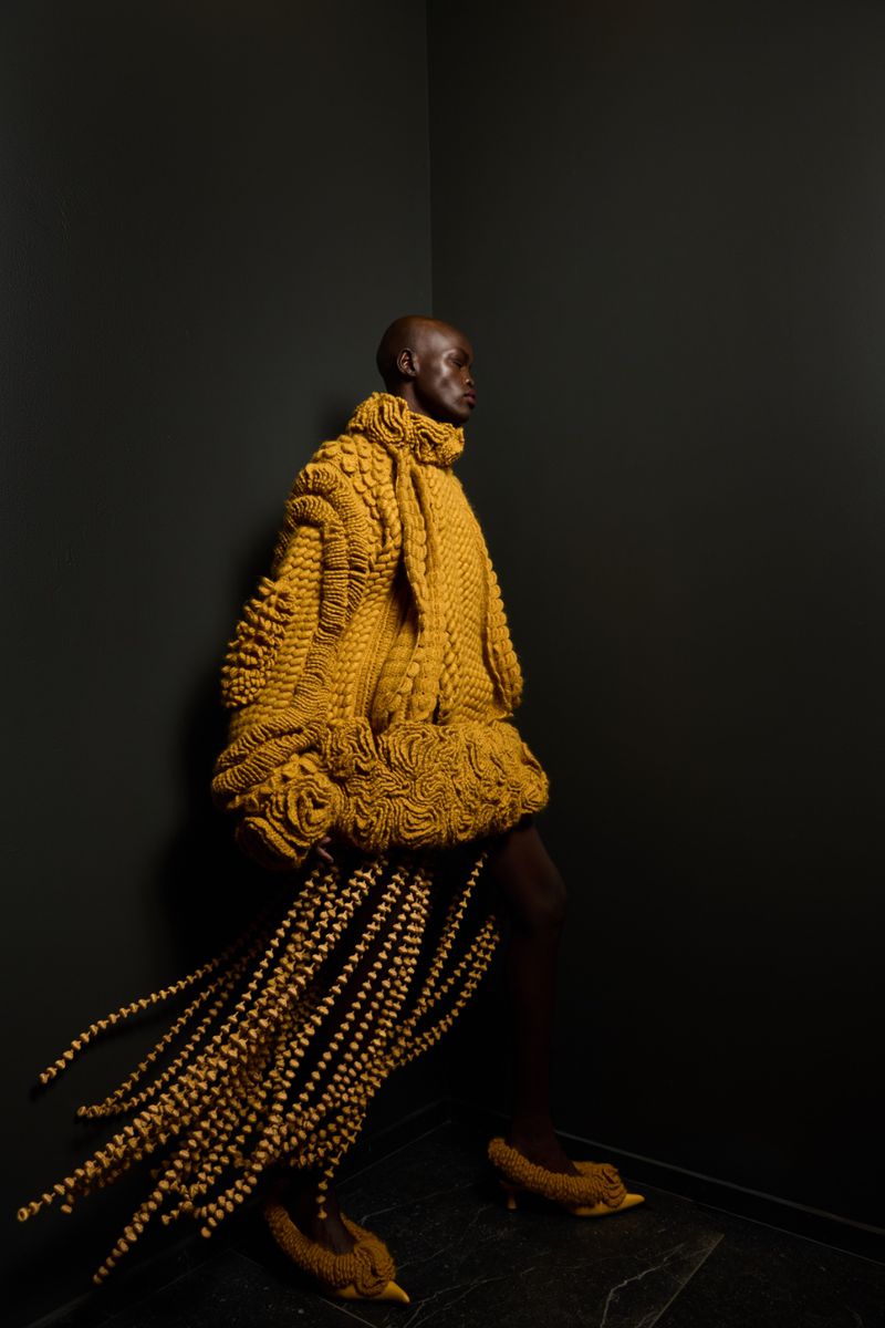 Bald adult fashion model in an elaborate yellow knit coat and shoes poses in a black corner photo studio with a dark floor.