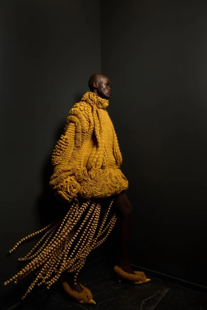 Bald adult fashion model in an elaborate yellow knit coat and shoes poses in a black corner photo studio with a dark floor.