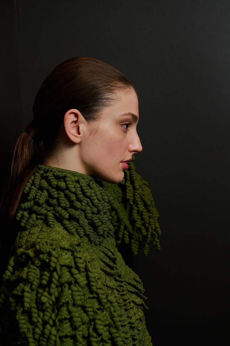 Profile view of a woman with auburn hair in a ponytail wearing an olive-green, textured sweater against a dark grey background.