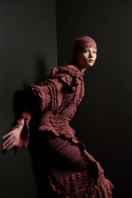 A person in a complex mauve knitted outfit poses in a studio against a dark background, fashion shot, detailed textures visible.