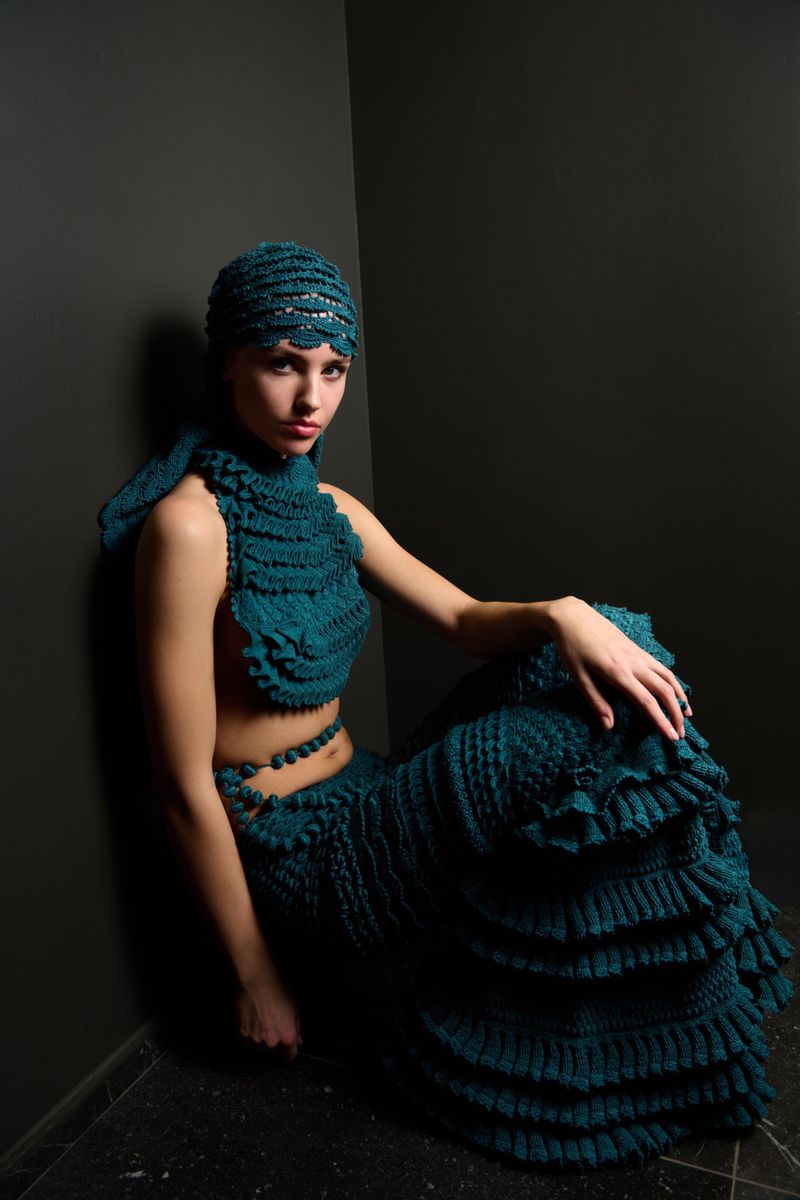 Woman poses indoors wearing teal crocheted top, skirt, and head covering. The scene is minimally lit against a dark grey wall.