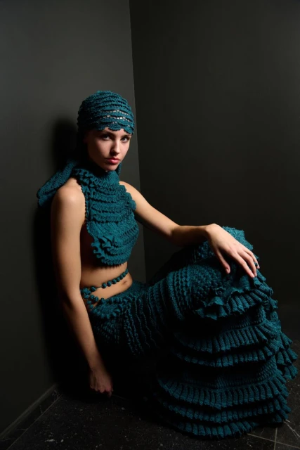 Woman poses indoors wearing teal crocheted top, skirt, and head covering. The scene is minimally lit against a dark grey wall.