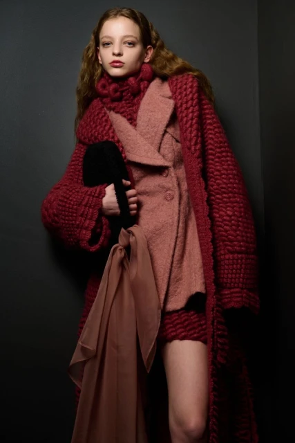 A model in a maroon knit coat and pink suit poses in front of a grey background, holding a black purse with a flowing brown scarf.