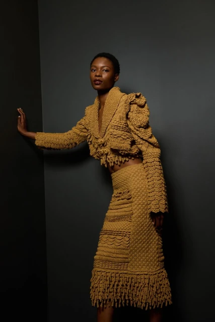 Portrait of a Black female fashion model wearing a golden yellow textured knit jacket and skirt with fringe details in a studio setting.