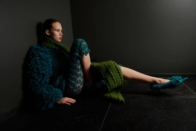 Model wearing textured teal knitwear and green scarf seated against a dark corner wall on tiled floor