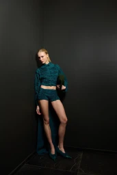 Model in textured teal matching jacket and shorts leaning against a dark grey wall holding a clutch