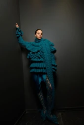 Model in textured teal knit sweater leaning against a dark grey corner wall in runway editorial