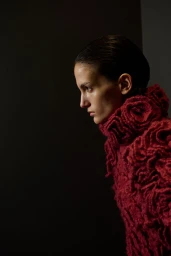 Model in profile wearing a textured red ruffled high-neck garment against a dark studio background