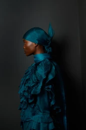 Model in profile wearing a teal ruffled blouse and matching headscarf against a dark studio backdrop