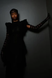 Model in black textured crochet dress and matching headpiece posing against a dark grey corner wall.