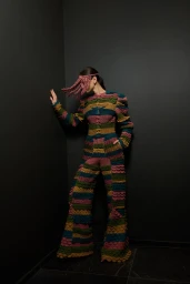 Model wearing a textured multi-colored knit jumpsuit and matching fringe headpiece standing against a dark grey corner wall.