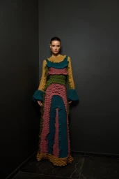 Model standing in a dark corner wearing a long textured knit dress with multi-colored patterns