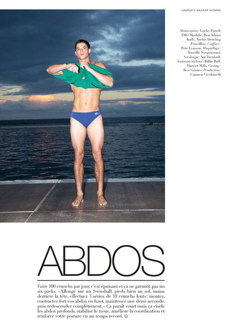 Athletic man in swim briefs and green shirt stands on a platform near water at dusk. Fitness fashion editorial outdoor shot.